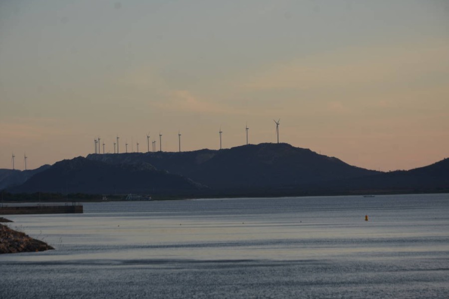 Grande espelho d’água com uma montanha ao fundo onde há aerogeradores de energia eólica sob a luz laranja do sol poente