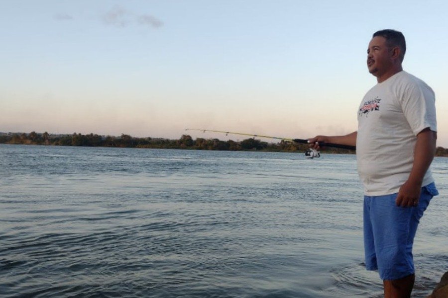 Homem de cabelos negro e bigode, usando uma camisa branca e um calção azul celeste enquanto pesca com uma varinha de pescar nas margens de um espelho d’água sob a luz do fim do dia