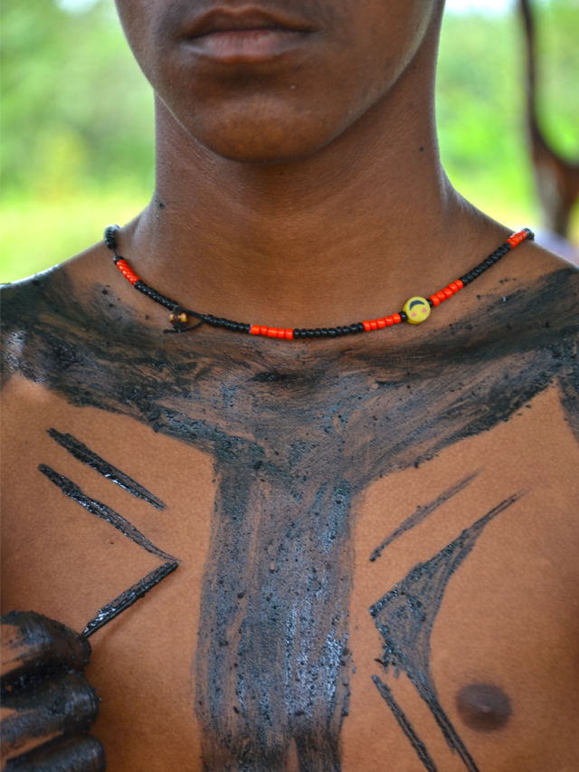 Fotografia do busto de um indígena de pele negra sendo pintado com pigmento preto pelas mãos de outro indígena. O que está sendo pintado usa um colar feito em sementes vermelhas e pretas