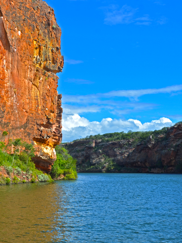 Fotografia de um grande paredão de rocha alaranjado coberto por vegetação às margens de um caudaloso espelho d´água sob céu azul