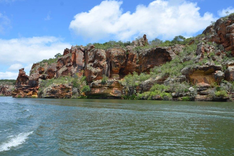 Grandes formações rochosas em tons alaranjados, repleta de vegetação entre as rochas que formam paredões margeando um grande rio de águas esverdeadas, sob céu azul com nuvens brancas