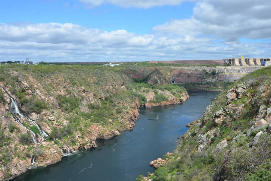 Foto de rio entre paredões rochosos com vegetação, uma pequena cachoeira descendo do lado esquerdo e uma represa ao fundo, sob céu azul com nuvens brancas