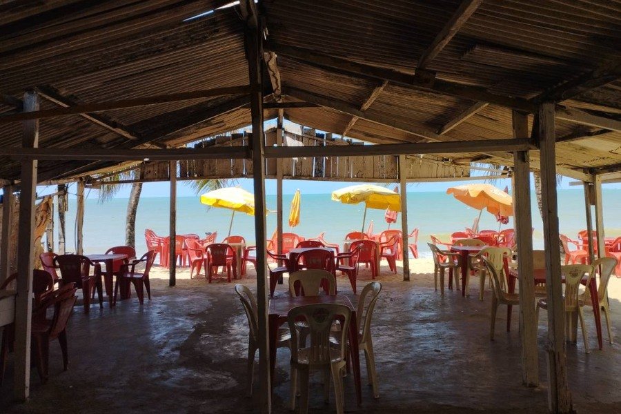 Conjunto de cadeiras e mesas de plástico na área de atendimento de um bar, à espera de visitantes. A maioria das cadeiras e mesas está no concreto, na sombra, sob a cobertura de um telhado. As demais estão na faixa de areia acompanhadas de guarda-sóis amarelos. Bem em frente está o azul do mar