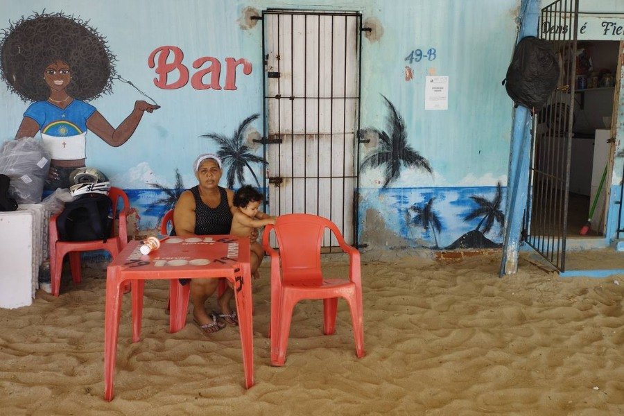 Na faixa de areia da praia, uma mulher negra com uma touca de cozinha está sentada numa cadeira plástica vermelha segurando uma bebê recém-nascida. Por trás da mulher, a fachada de uma bar, cuja porta está aberta. A parede do bar tem pintura na cor azul e a frase “Deus é Fiel” escrita. Também tem coqueiros desenhados e uma mulher negra de cabelo afro com uma camisa semelhante à bandeira do Estado de Pernambuco