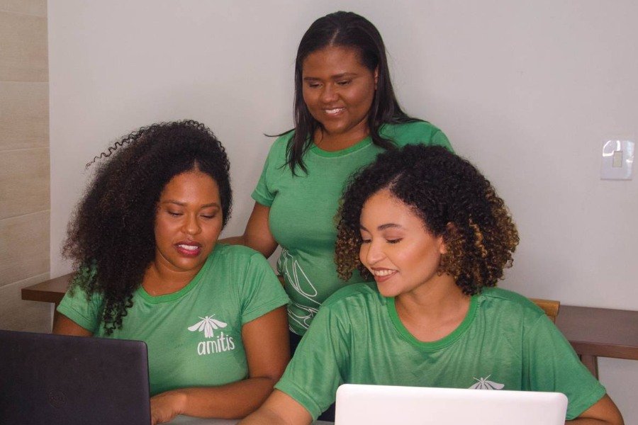Três mulheres negras usam camisetas verdes. Uma, de cabelos lisos, está em em pé entre as outras duas que estão sentadas à frente de computadores. As outras duas têm capelos crespos