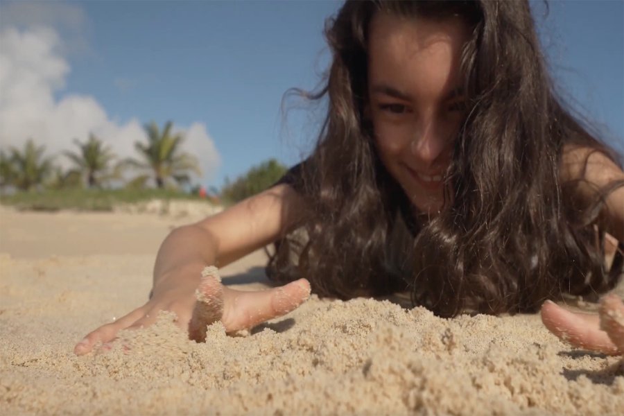 Menina de cabelos castanhos escuros e cumpridos deitada na praia enquanto brinca com areia sob o céu azul
