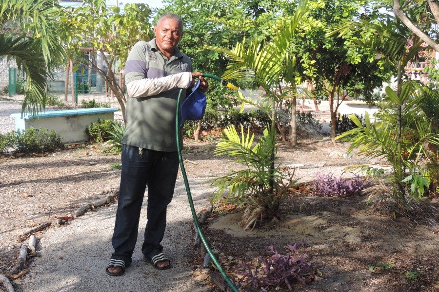 Homem negro de de meia idade veste calça jeans e blusa verde com mangas cinza. Segura uma mangueira com a qual agoa plantas próximas a ele em um parque.