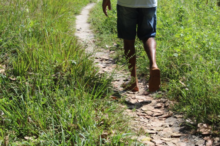 Na foto, são mostradas as pernas e cintura de um homem indígena caminhando sob uma estradinha de barro, ao redor, pequenos matinhos