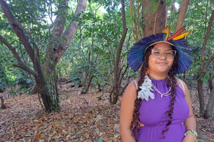 Jovem indígena usa um cocar feito em penas de aves azuis, amarelas e vermelhas. Ela tem os cabelos longos e trançados e usas óculos de grau e vestido roxo