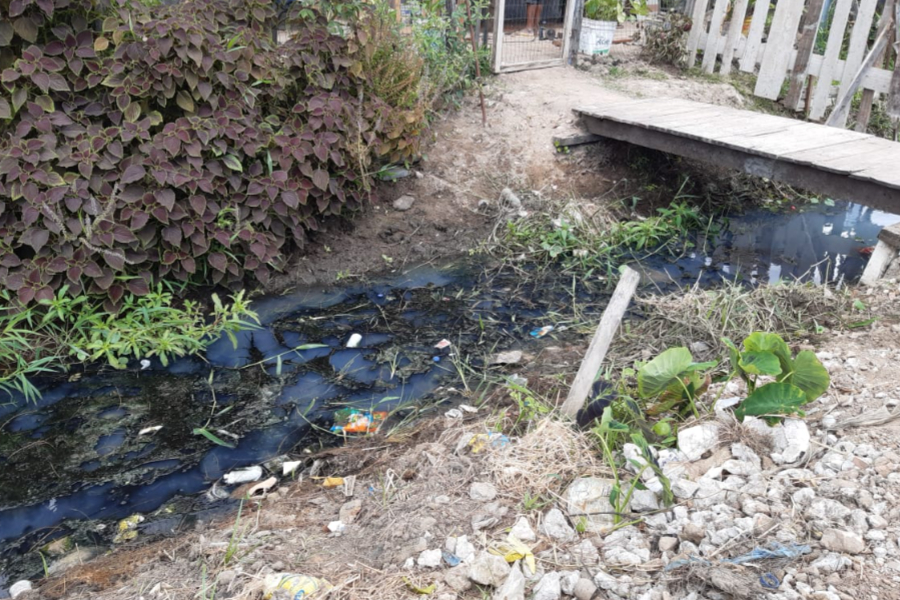 Canal com esgoto e resíduos sólidos. Nas laterais há vegetação rasteira e pedras. Em segundo plano há uma pequena ponte de madeira