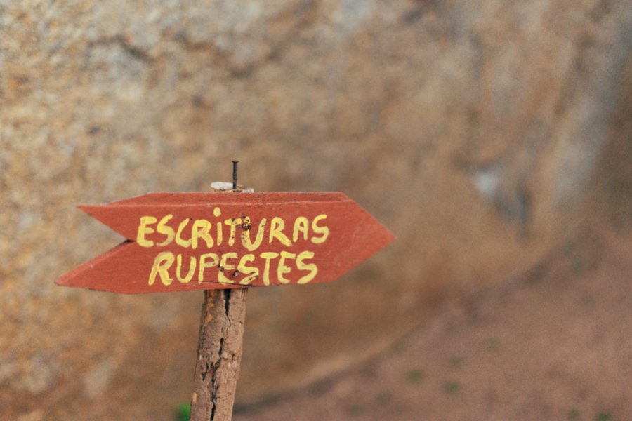 Placa vermelha em formato de seta onde se lê, em letras amarelas, "ESCRITURAS RUPESTRES". Ao fundo, uma formação rochosa avermelhada desfocada