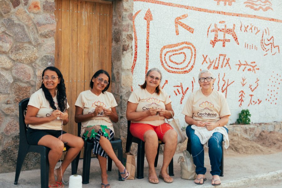 Quatro mulheres sentadas em cadeiras na calçada sorriem para a câmera enquanto fazem crochê