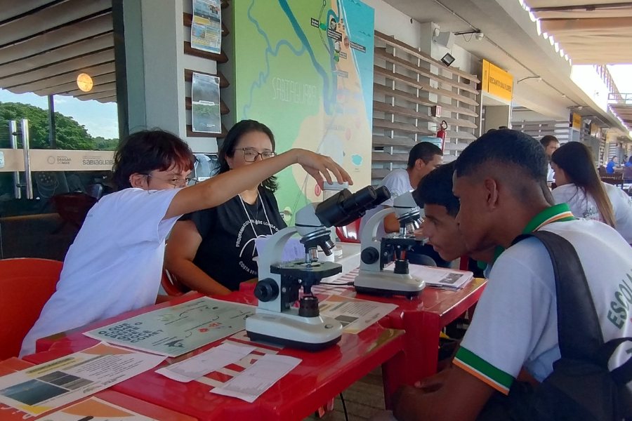 Dois estudantes negros sentados em frente a uma mesa onde há microscópios. Do outro lado da mesa há duas mulheres expondo o material e auxiliando no uso dos equipamentos