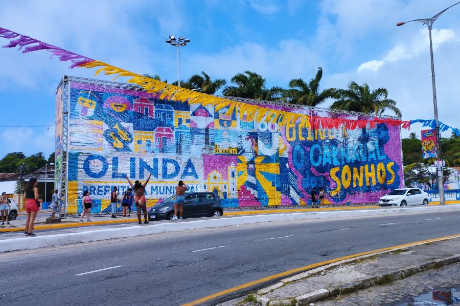 Do outro lado de uma avenida, painel gigante, onde predominam as cores azul e amarelo destaca OLINDA, PREFEITURA MUNICIPAL, OLINDA O CARNAVAL DOS SONHOS. Por trás é possível observar a ponta da copa de coqueiros, acima céu azul com nuvens e à frente algumas pessoas observam e fotografam e um carro escuro e um branco passam