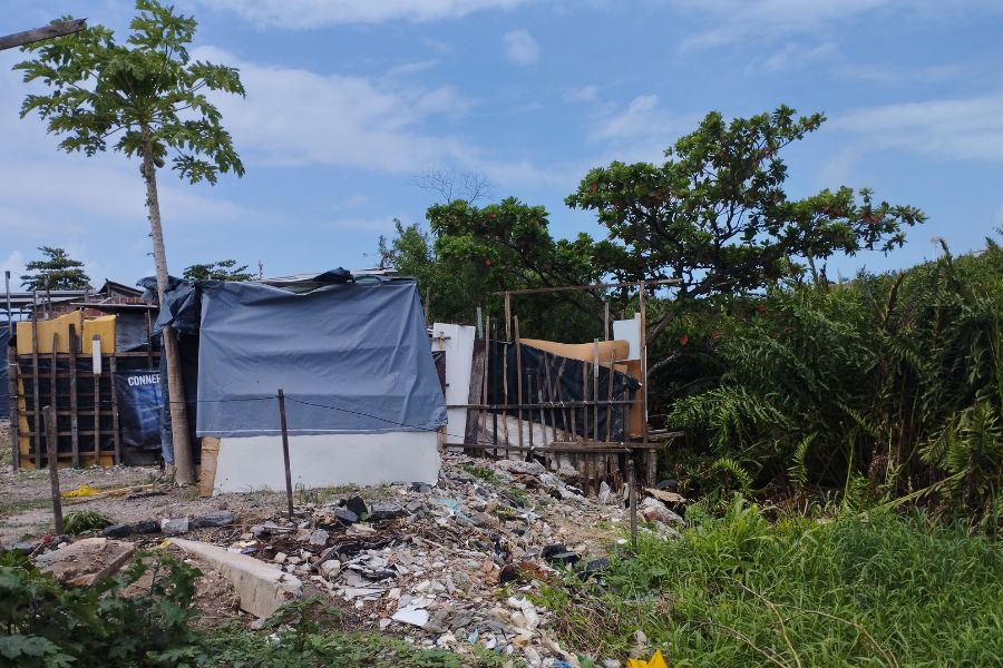 Sobre entulhos, ao lado de vegetação, erguem-se barracos sob céu azul com nuvens brancas