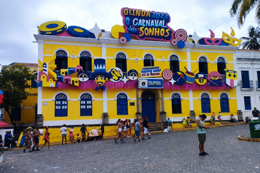 Prédio histórico pintado de amarelo, com detalhes brancos, portas e janelas azuiscom topo em arcoenfeitado com motivos em alusão ao Carnaval pernambucano com os dizeres OLINDA O CARNAVAL DOS SONHOS. Pessoas caminha na rua de calçamento em frente