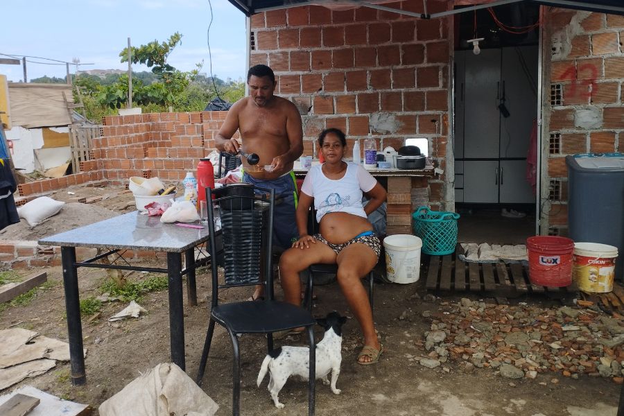 Em frente a uma construção de tijolos, uma mulher grávida está sentada rm uma cadeira ao lado de uma mesa olhando para a câmera, enquanto um homem em pé serve um prato. À frente da mulher, um cachorro branco e preto olha para ela