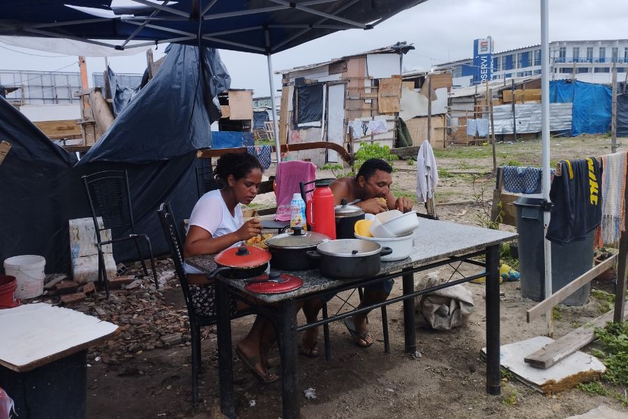 Mulher e homem comem sentados atrás de uma mesa onde há panelas, sob um toldo azul rasgado com barracos ao fundo