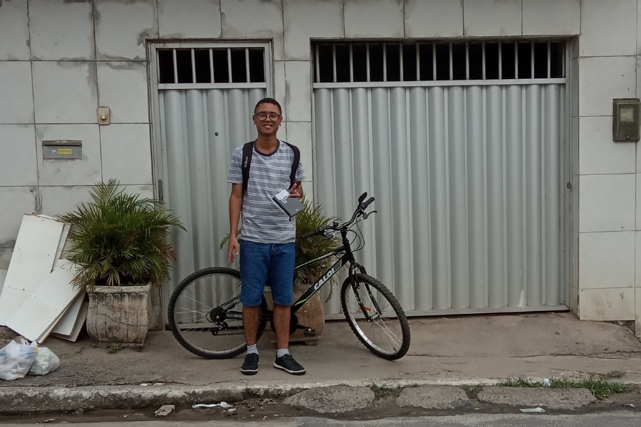 No centro da imagem, um homem pardo, jovem, de óculos. Ele está em pé na calçada, de frente, com uma mochila nas costas, um caderno na mão, e na companhia de uma bicicleta preta. Atrás dele, uma casa com cerâmica branca