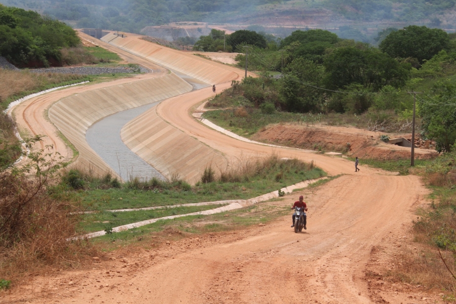 Canal feito com cimento e com pouca água serpenteia terra avermelhada com vegetação verde dos dois lados. Na margem direita um homem de camisa vermelha pilota uma motocicleta. Mais adiante é possível ver um transeunte a pé