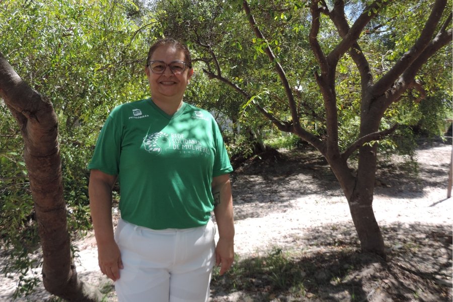 Foto de mulher branca com os cabelos presos. Ela usa óculos, uma camiseta verde e uma calça branca e está entre árvores