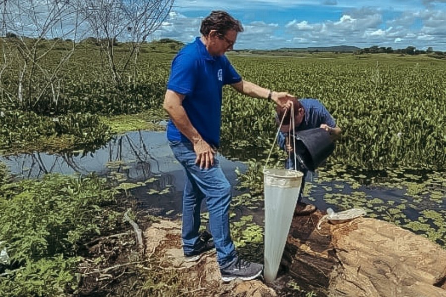 Homem em pé em coleta de amostra de água segura saco que filtra a amostra. Outro homem o ajuda com um balde. Eles estão em uma rocha na margem de um manancial rio coberto por aguapés