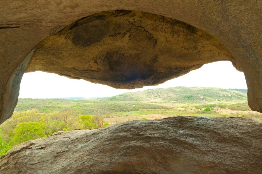 Em primeiro plano as paredes da caverna formam o desenho de um chapéu de cangaceiro. Ao fundo, a paisagem da Serra do Teixeira