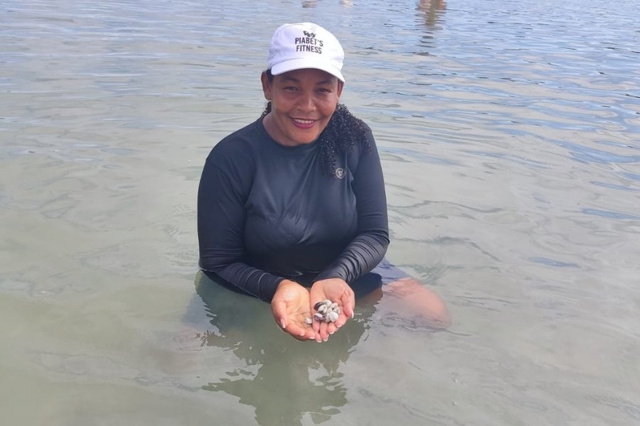 Uma mulher negra segura mariscos num estuário (transição do rio com o mar). Ela veste uma camisa UV preta e um boné branco. Ao fundo, próximo ao mangue, outras mulheres também coletam mariscos
