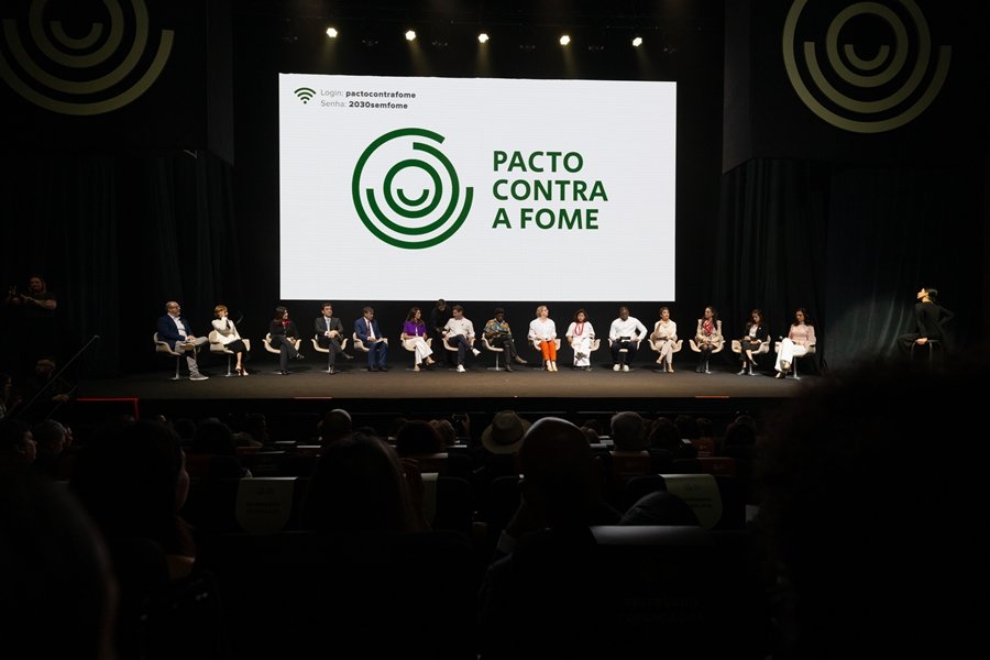 Auditório escuro, com um telão branco onde há dizeres em verde: "Pacto contra a fome". No palco do auditório há pessoas sentadas em cadeiras