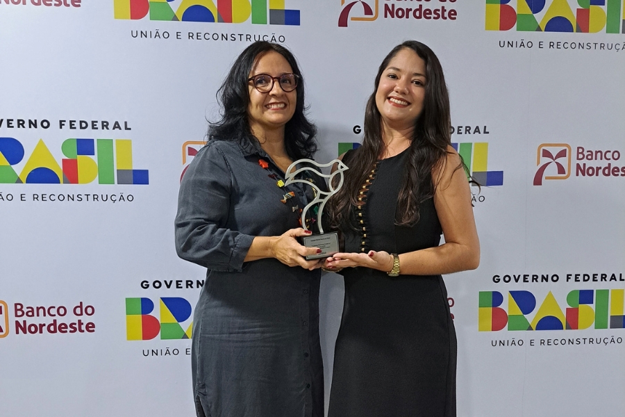 Duas mulheres morenas, uma de cabelos mais curtos e óculos, outra de cabelos longos, vestidas de preto, seguram um troféu de metal em formato de pássaro