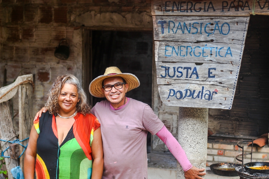 Casal sorri abraçado. Ela é parda e tem o cabelo grisalho nas raizes e avermelhado e mais cacheado nas pontas e usa roupa colorida. Ele usa chapéu de palha, óculos e camiseta UV rosa sobreposta por camiseta meia manga rosa de outro tom. Eles estão em frente a uma construção de tijolo com uma placa de madeira com os dizeres: "ENERGIA PARA VIDA: TRANSIÇÃO ENERGÉTICA JUSTA E popular"