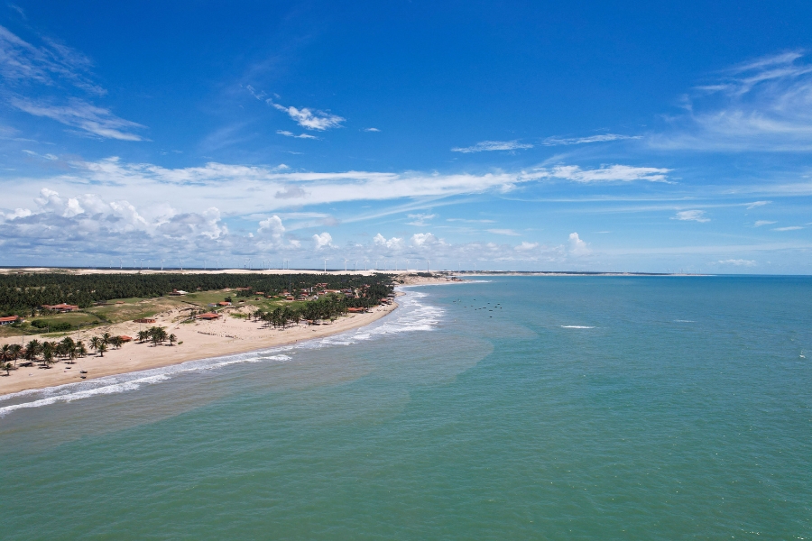 Foto aérea do mar verde com faixa de areia no horizonte, no meio da imagem, à esquerda com vegetação e pequenas construções. Tudo sob céu azul com poucas nuvens brancas