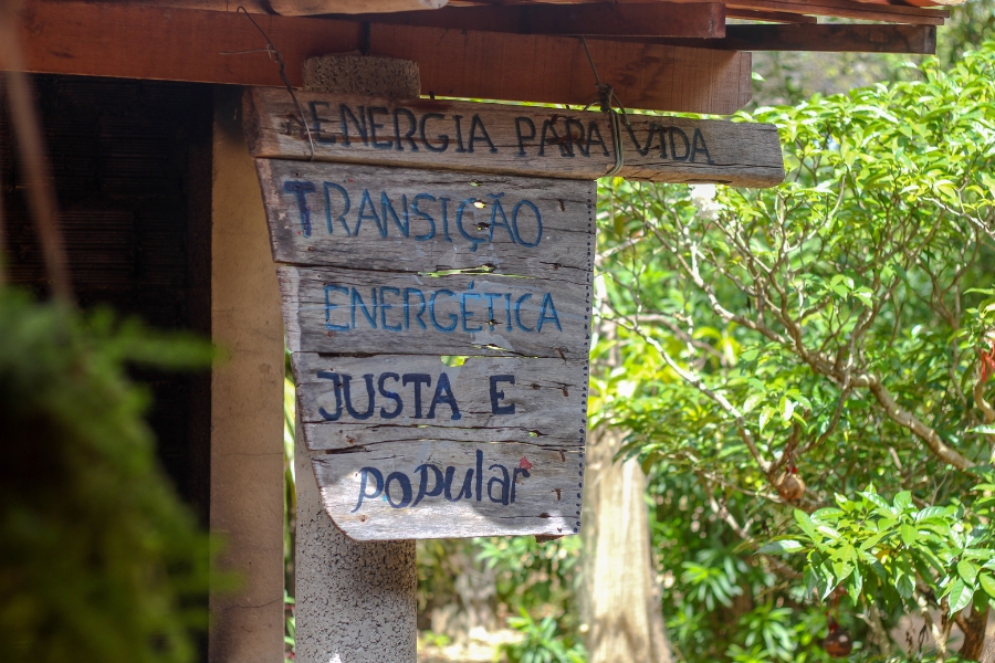 Placa de madeira com os dizeres: "ENERGIA PARA VIDA: TRANSIÇÃO ENERGÉTICA JUSTA E popular". Ao fundo vegetação desfocada