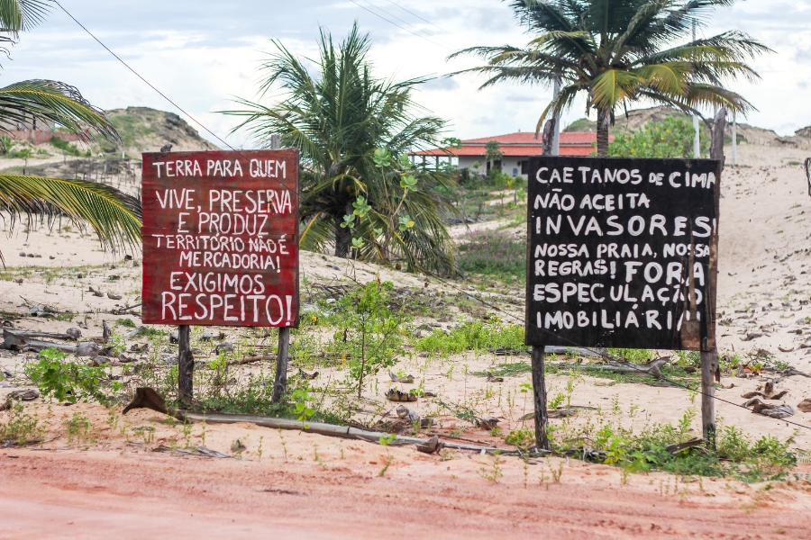 Terreno arenoso com coqueiros e casa ao fundo apresenta duas placas, uma de fundo vermelho onde lê-se "TERRA PARA QUEM VIVE, PRESERVA E PRODUZ. TERRITÓRIO NÃO É MERCADORIA! EXIGIMOS RESPEITO!", outra de fundo preto: "CAETANOS DE CIMA NÃO ACEITA INVASORES! NOSSA PRAIA, NOSSAS REGRAS! FORA ESPECULAÇÃO IMOBILIÁRIA"