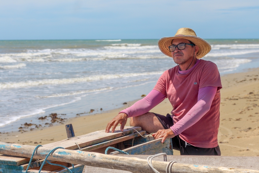 Homem vestindo camisa UV rosa sob camiseta meia manga rosa em outro tom, calção preto, chapéu de palha e óculos olha em frente apoiado em pequena embarcação de madeira na areia da praia tendo o mar ao fundo sob céu azul
