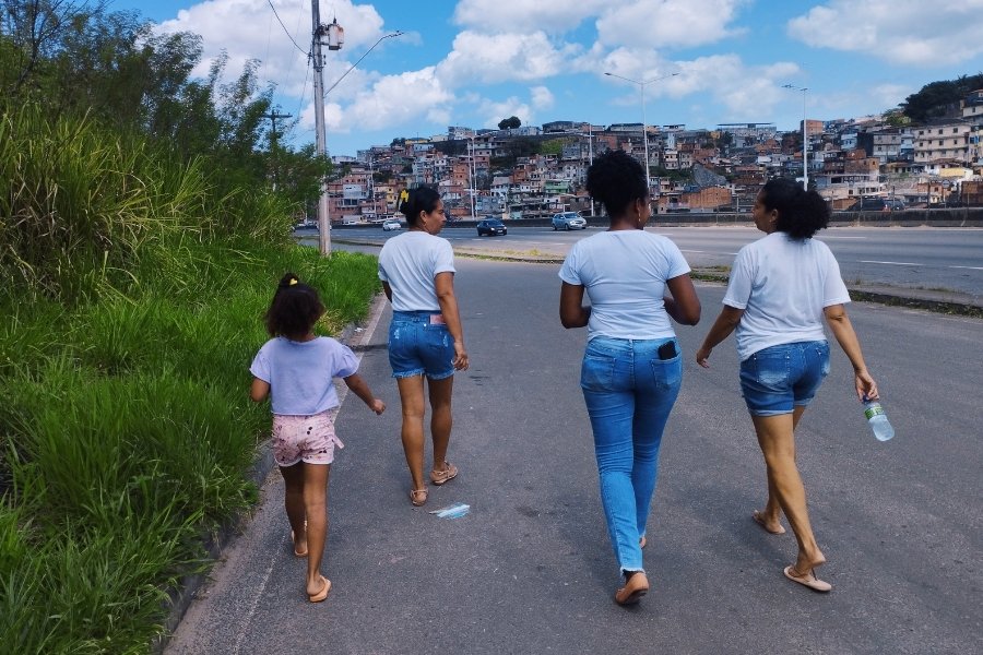 Vistas de costa, três mulheres adultas e uma criança caminham às margens de uma avenida expressa. À esquerda delas, um mato alto. Na direita, um fluxo suave de carros e uma comunidade em área de morro com grande adensamento populacional