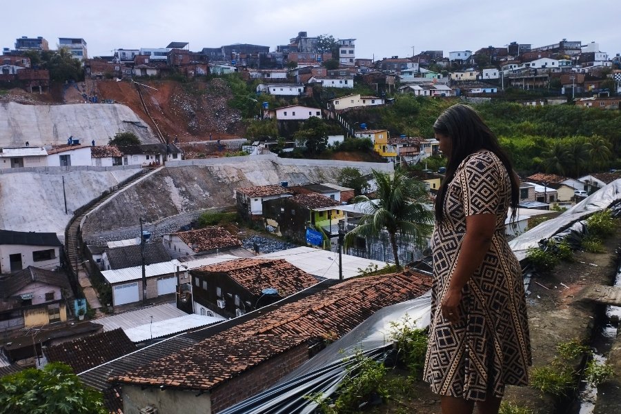 Na parte alta de uma favela, uma mulher negra, de cabelos longos, observa no horizonte o morro vizinho. Ela está na frente de uma casa, na beira de uma encosta coberta com lona plástica preta