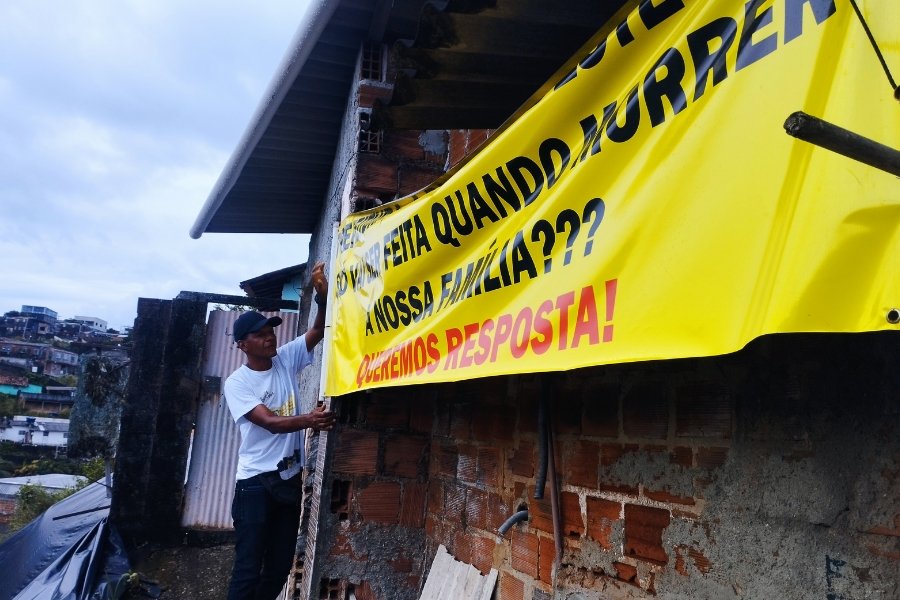 Um homem negro, de calça, camisa e boné, observa o bandeirão amarelo, com uma mensagem crítica, posicionado na porta de uma casa. À sua esquerda, uma encosta coberta com lona plástica preta