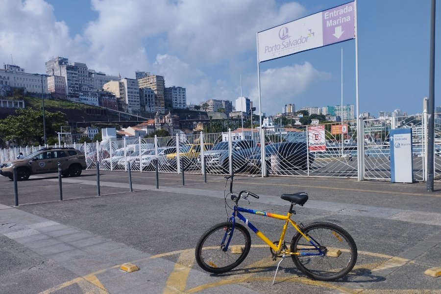 Em um dia ensolarado, no centro da imagem, uma bicicleta azul e amarela parada em um estacionamento, próximo uma marina com embarcações. Ao fundo, construções antigas no topo de uma colina