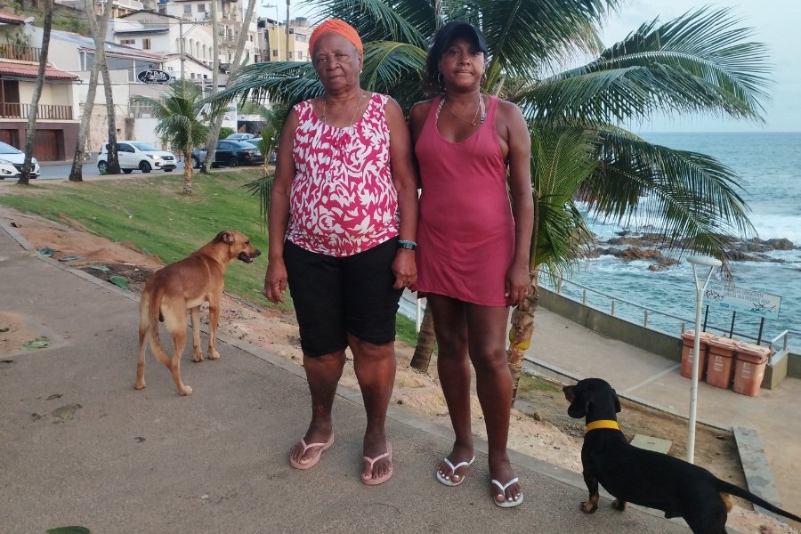 Duas mulheres negras olham de frente para a câmera. A da direita é adulta, usa vestido vermelho e boné preto. A outra é idosa, e usa um lenço laranja na cabeça. Ao lado delas, estão dois cachorros, um preto e outro amarelo. Ao fundo se vê uma praia com rochas, um coqueiro e construções à beira-mar