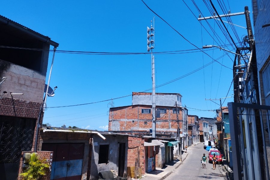 Céu azul e muito sol sobre uma rua estreita, dentro de uma comunidade periférica A rua tem construções simples, fiações aéreas e um poste com megafones que serve como sirene sonora para os moradores