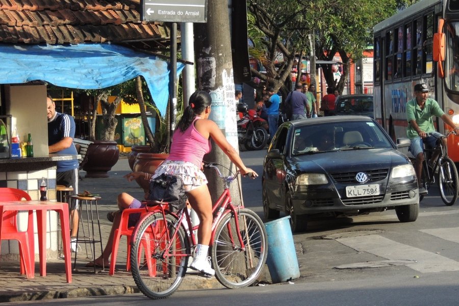 Uma mulher negra veste blusa rosa e saia branca e pedala uma bicicleta simples. Ela está em um cruzamento, esperando o sinal abrir, entre duas vias movimentadas. À sua direita, passam um carro, um ônibus e outra bicicleta