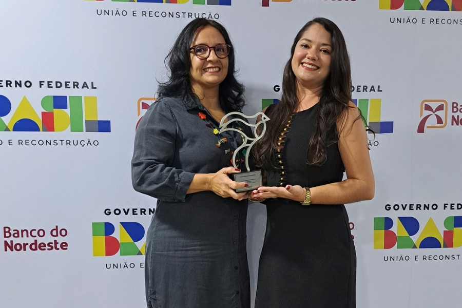 Duas mulheres morenas, uma de cabelos mais curtos e óculos, outra de cabelos longos, vestidas de preto, seguram um troféu de metal em formato de pássaro