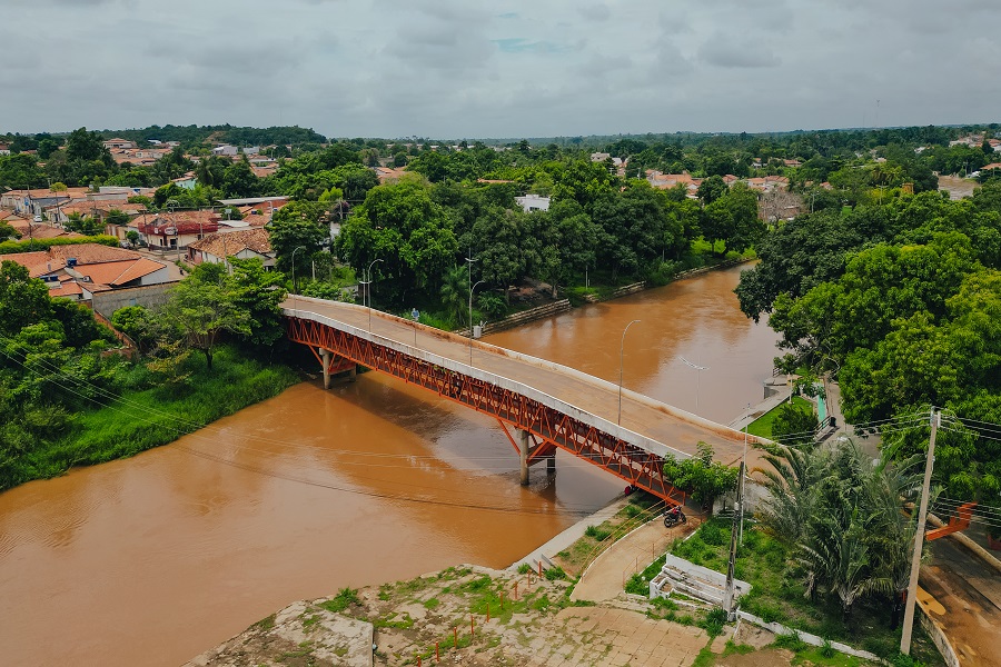 Foto aérea panorâmica colorida de uma ponte sobre rio barrento e nas margens vegetação e moradias. Ao fundo, céu nublado