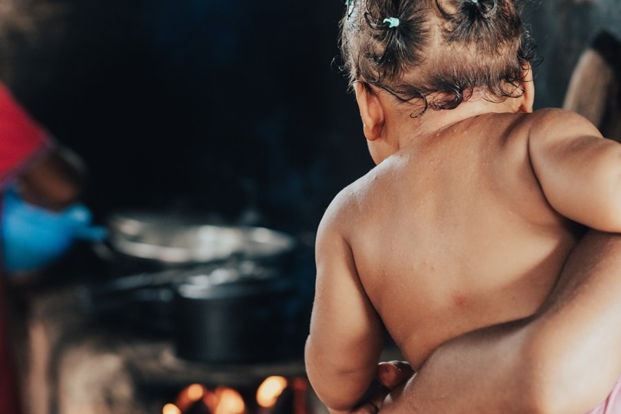 Foto colorida mostra um bebê de pele negra de costas sendo segurado pelos braços de um adulto não identificado, ao fundo há panelas em um fogão à lenha