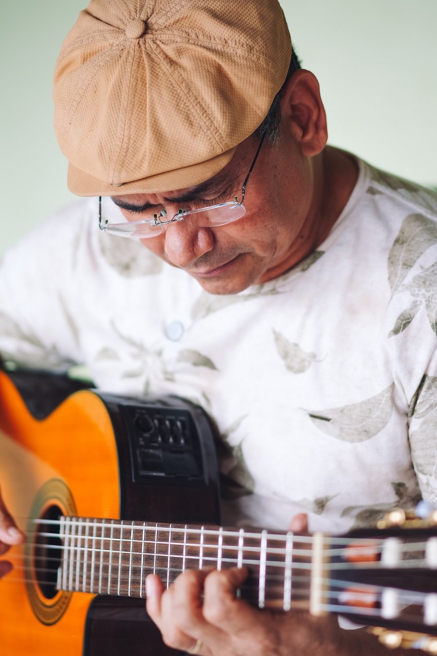 Imagem colorida de homem moreno, de boné e óculos, olhando para o violão enquanto toca o instrumento