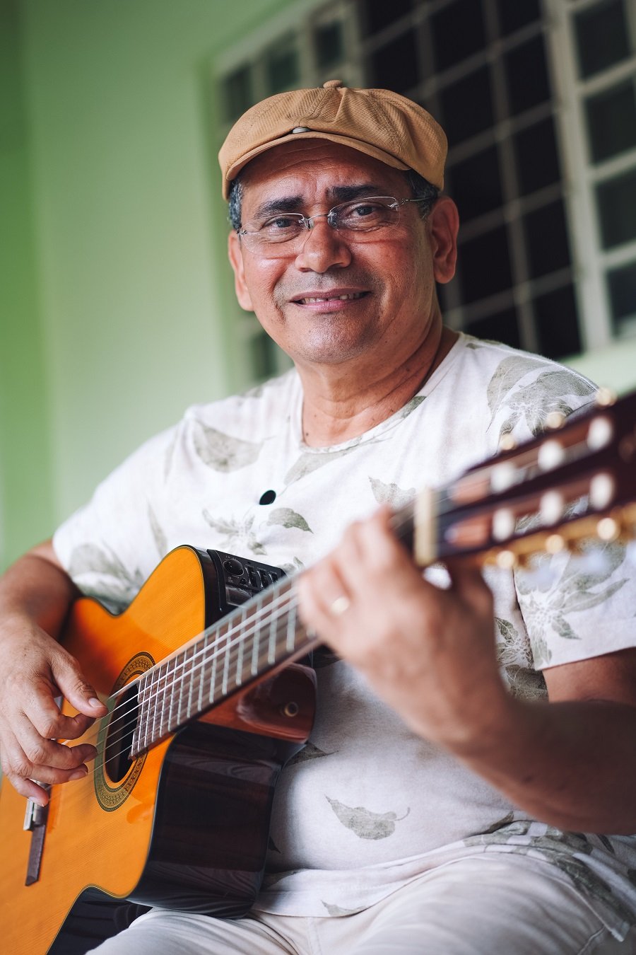 Imagem de homem moreno de boné e óculos, sentado, sorrindo para a foto e tocando violão