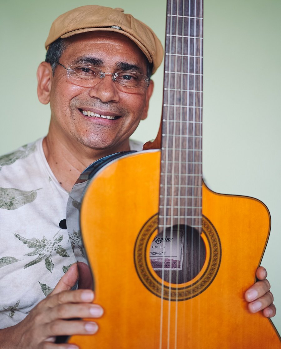 Foto colorida de homem moreno de boné e óculos sorrindo e segurando um violão em pé