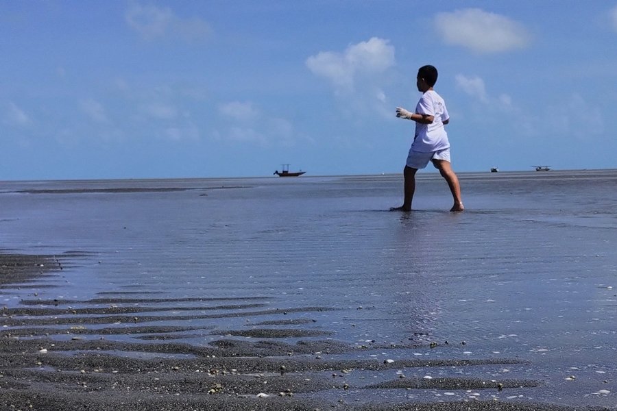Foto de um menino, vestido de calção azul e camisa branca, caminhando pela praia sob um céu azul