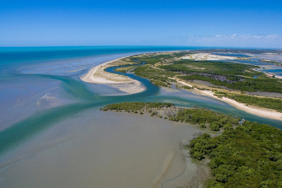 Imagem aérea de uma faixa de praia, com mar azul, encontro de um rio com mar e vegetação no entorno da praia